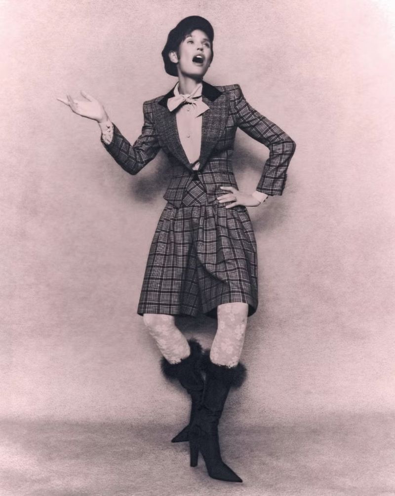 A full shot of a stylish woman in a plaid suit, textured tights, and heeled boots against a neutral backdrop, posing expressively.