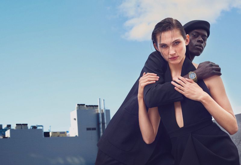 Editorial fashion shot of an African-descent man and Caucasian woman in dark clothing against an urban backdrop under a blue sky.