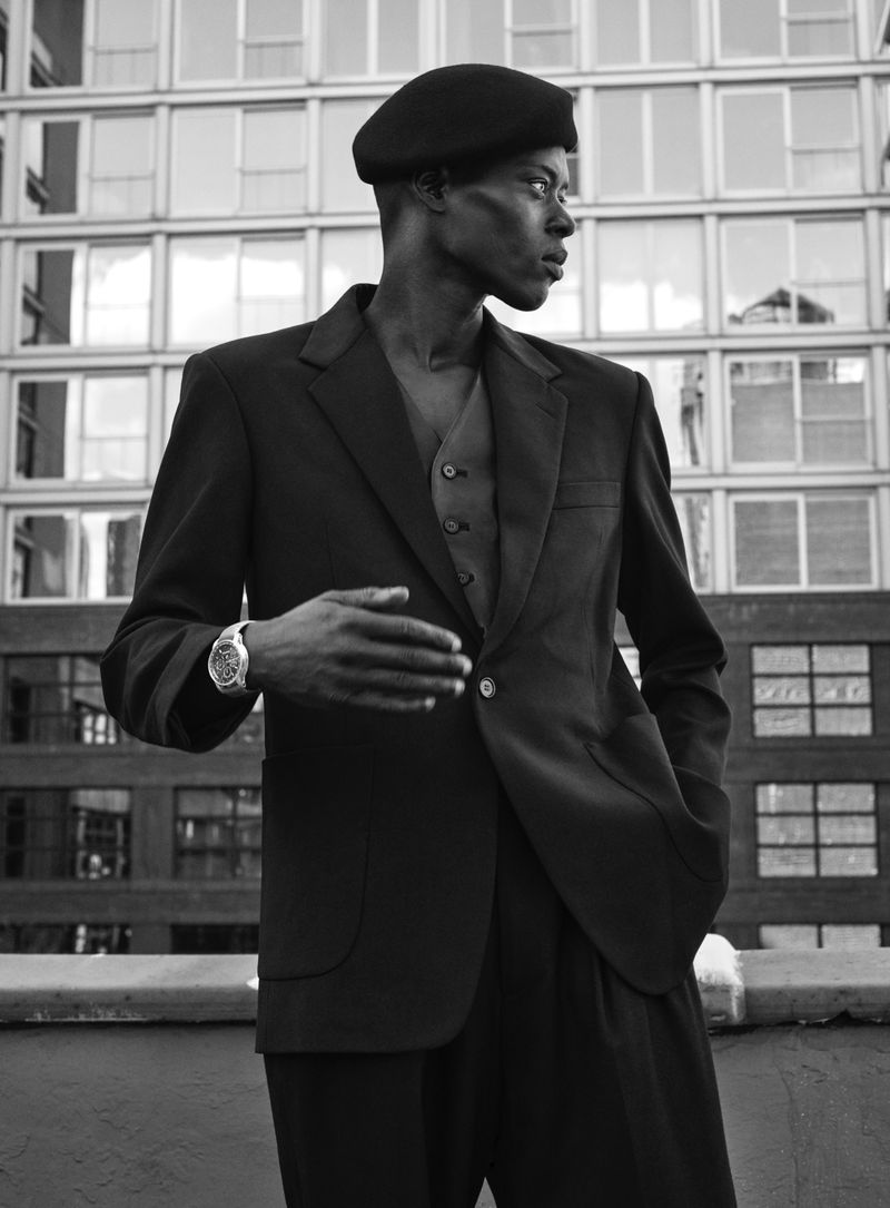 Stylish African-American male model in a black suit and beret poses against a modern building for a fashion editorial shoot.