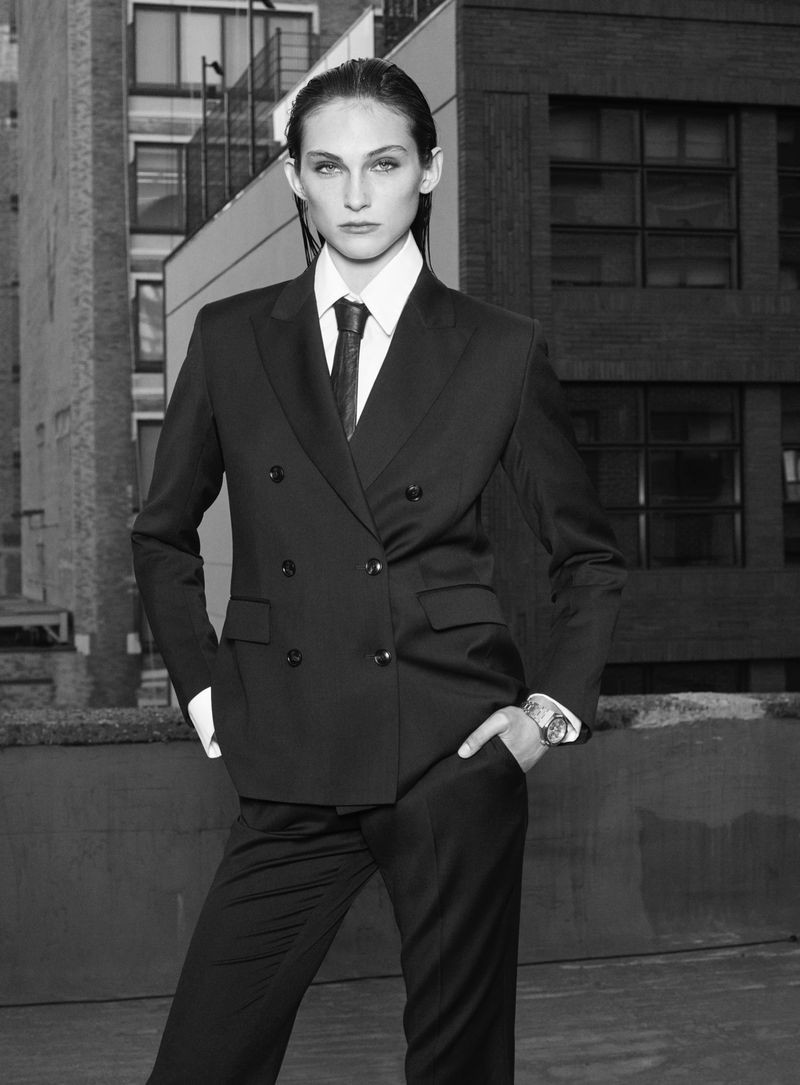 Adult fashion model in a black double-breasted suit, white shirt, and black tie poses against a backdrop of urban architecture.