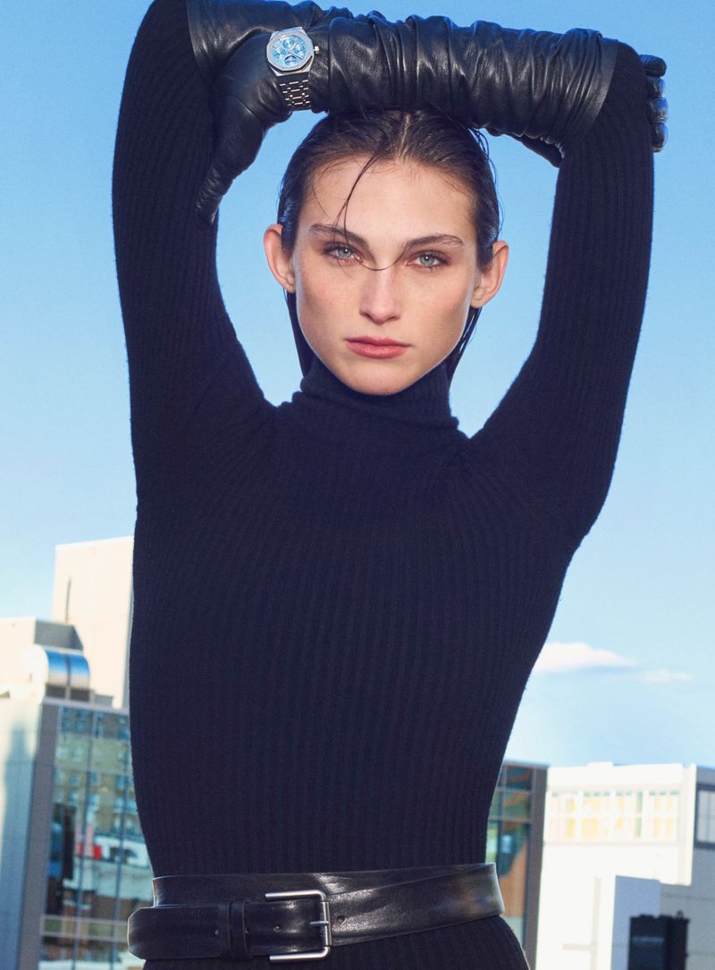 A woman poses outdoors wearing a black turtleneck dress, gloves, and a watch, set against a cityscape and blue sky, captured in a medium shot.