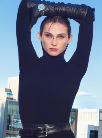 A woman poses outdoors wearing a black turtleneck dress, gloves, and a watch, set against a cityscape and blue sky, captured in a medium shot.