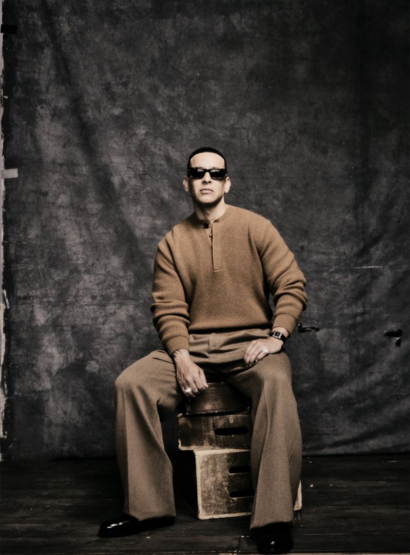 Man in sunglasses poses in front of a mottled gray background, sitting on a wooden crate wearing a brown sweater and tan trousers.