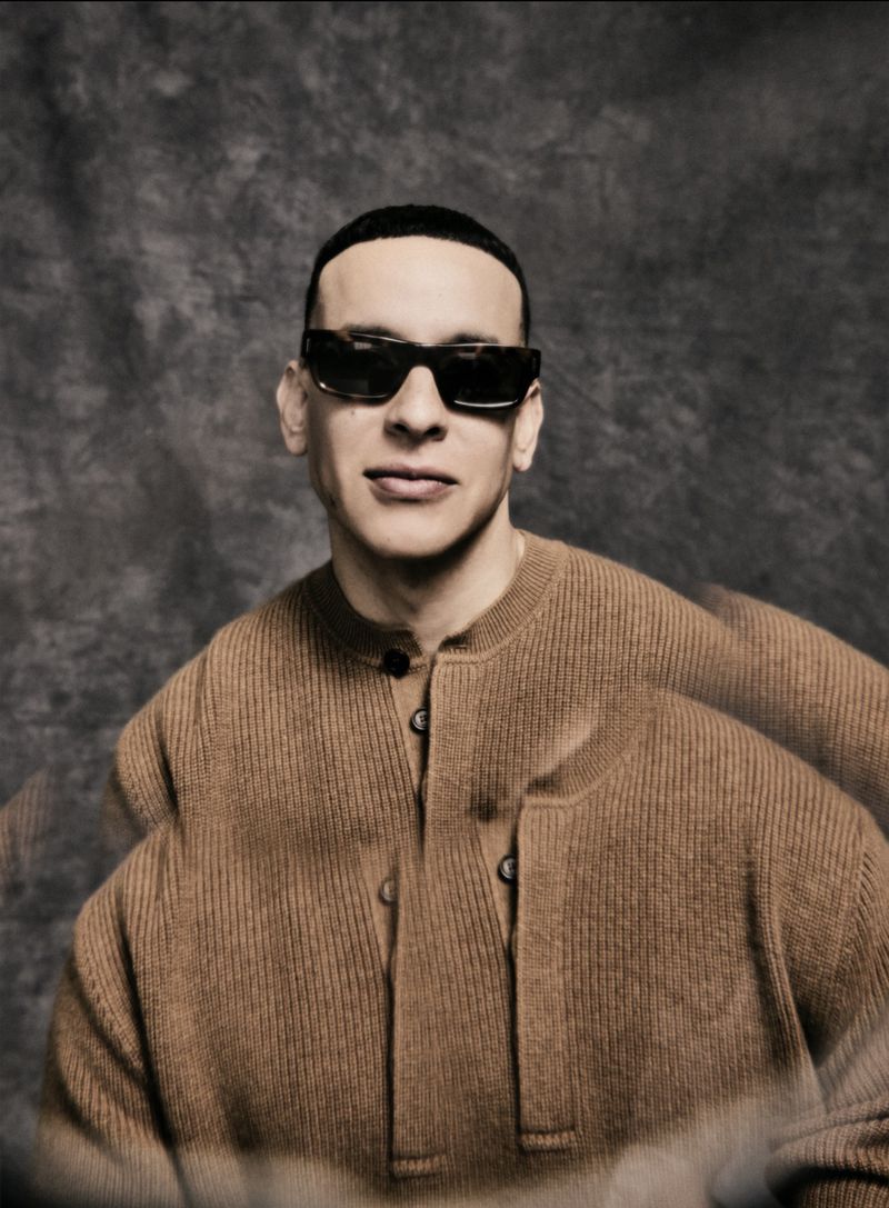 Daddy Yankee wearing dark sunglasses and a ribbed brown knit sweater against a mottled grey textured studio backdrop.