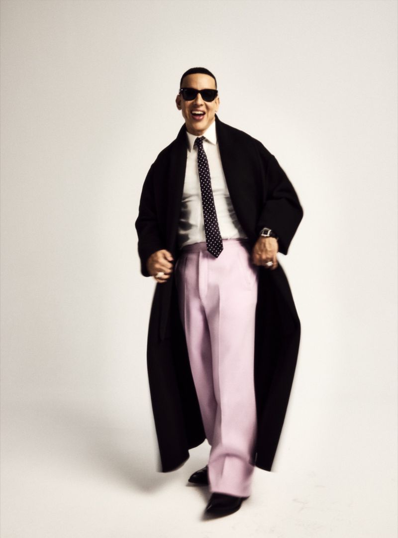 Daddy Yankee posing in a long black coat, white dress shirt, polka dot tie, and wide-leg pink trousers against a white backdrop.
