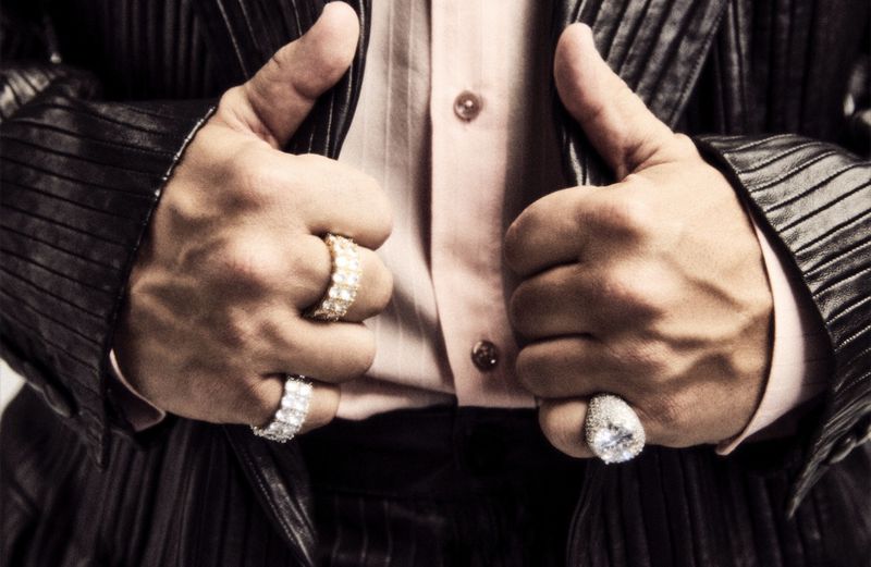 Close-up on a person's hands, adorned with prominent gold and diamond rings, giving a thumbs-up gesture, wearing a pink shirt and black jacket.