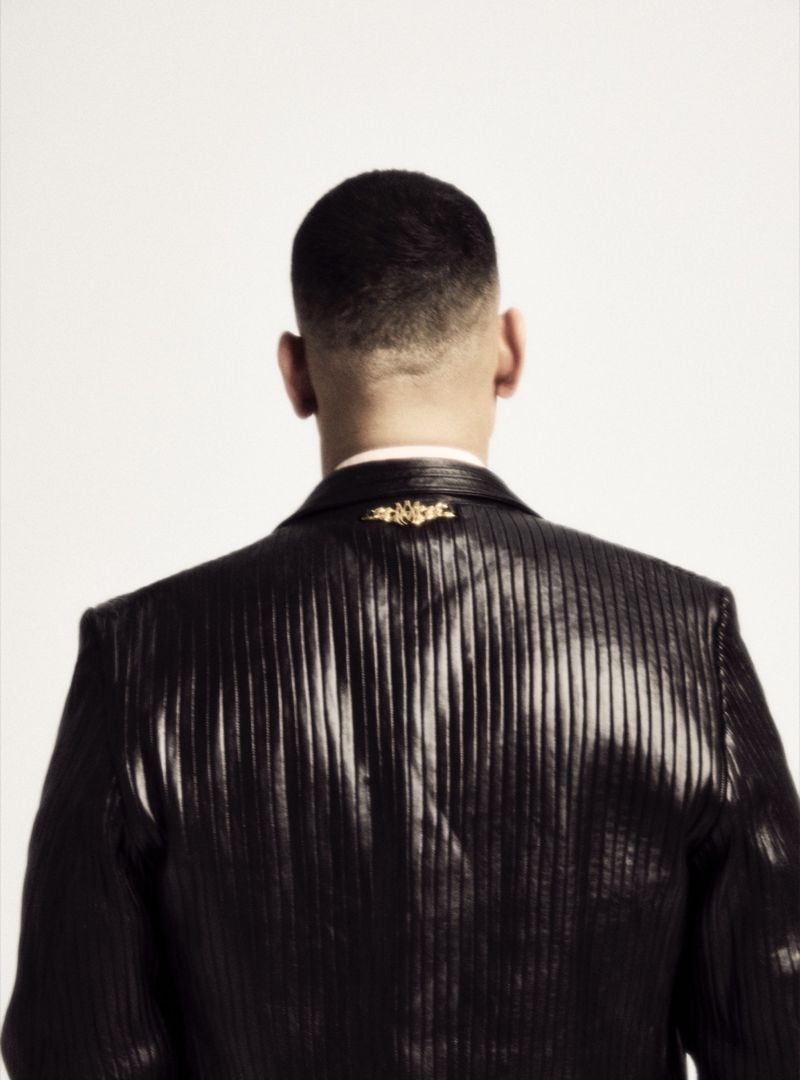 Back view of a stylish adult male with a short haircut wearing a textured black suit jacket with an ornate gold brooch against a white background.