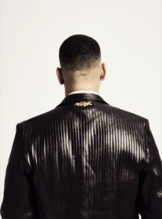 Rear view of a man with a short fade haircut wearing a glossy pleated black leather jacket against a plain white background.