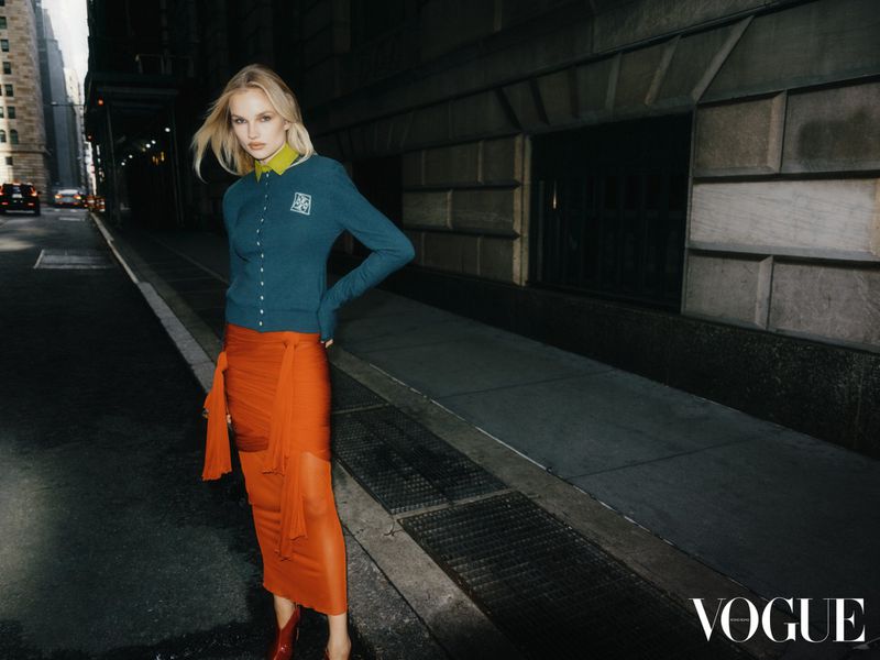 Blonde fashion model poses on an urban city street wearing a teal cardigan and sheer orange skirt with a Vogue logo in the corner.