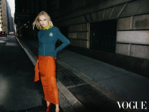 Blonde fashion model poses on an urban city street wearing a teal cardigan and sheer orange skirt with a Vogue logo in the corner.