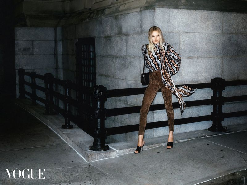 Fashion shot of a blonde adult woman posing in brown pants and a patterned blouse in an urban outdoor setting with grey stone walls.