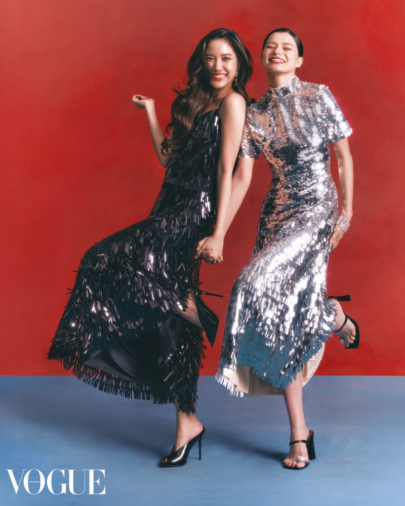 Two smiling women pose in sparkling gowns against a vibrant red backdrop, showcasing high fashion and elegance for a Vogue editorial.