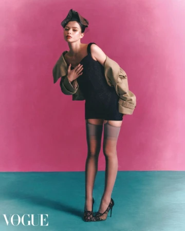 Full-length portrait of a model in a black mini dress, beige trench coat, and stockings posing against a bright pink studio backdrop.