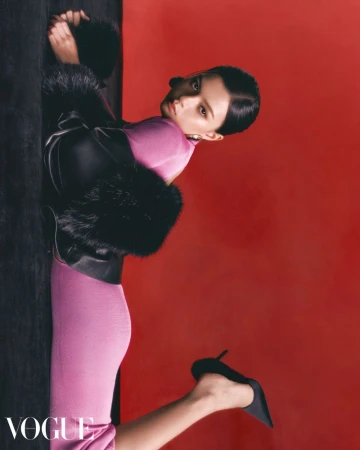Fashion editorial featuring a woman in a pink dress and fur-trimmed coat, reclining against a dark object with a red studio background.