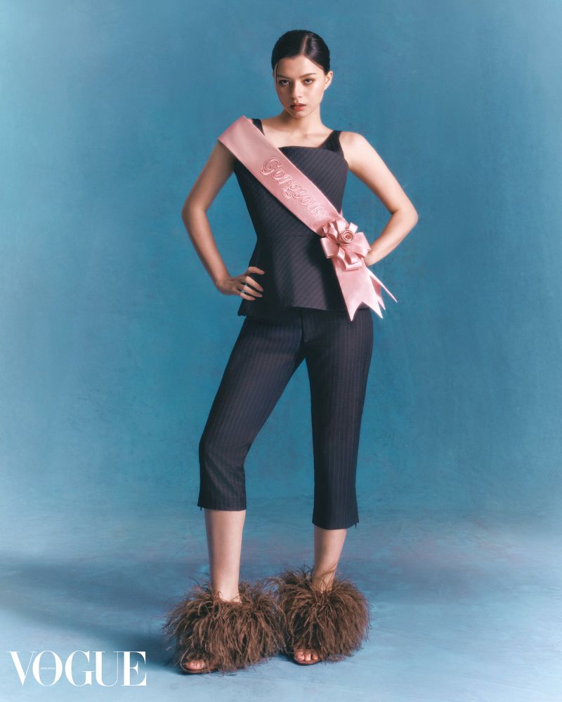 A woman in a black top and cropped pants poses in front of a blue background wearing feathered shoes and a pink sash. Fashion editorial.