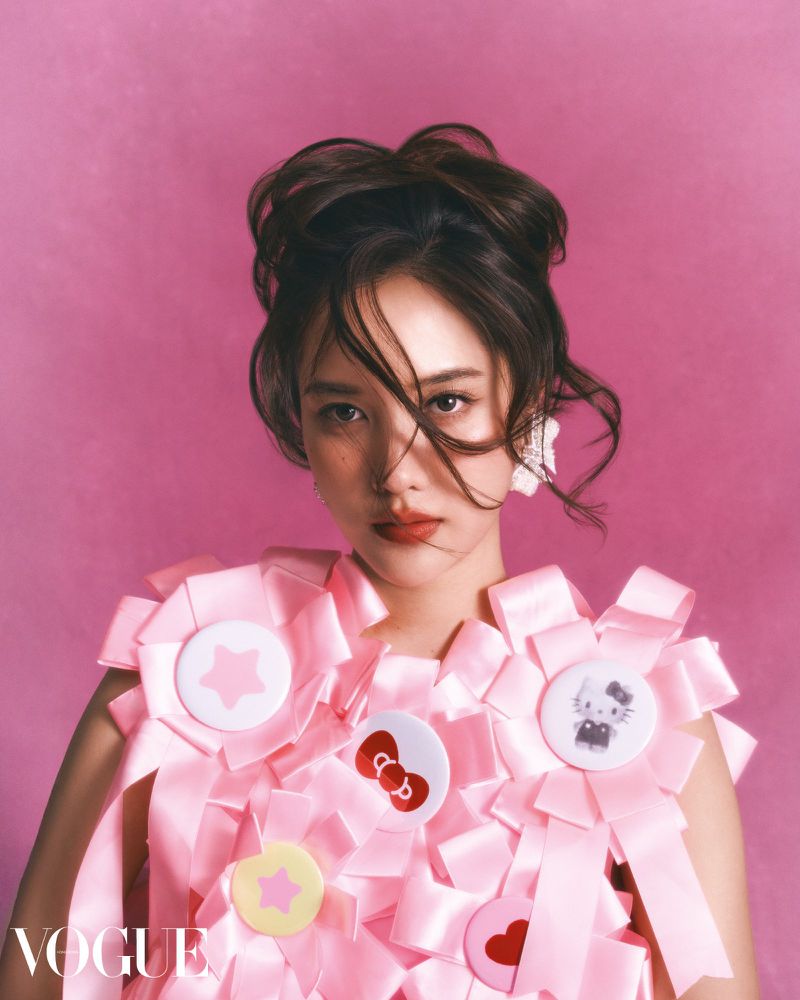 Portrait of a young woman with brown hair in an updo, wearing a pink ribbon and badge dress against a bright pink backdrop.