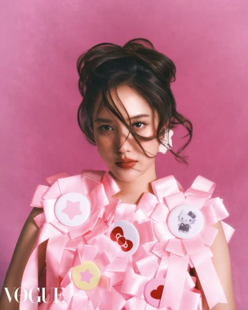 Portrait of a young woman with brown hair in an updo, wearing a pink ribbon and badge dress against a bright pink backdrop.