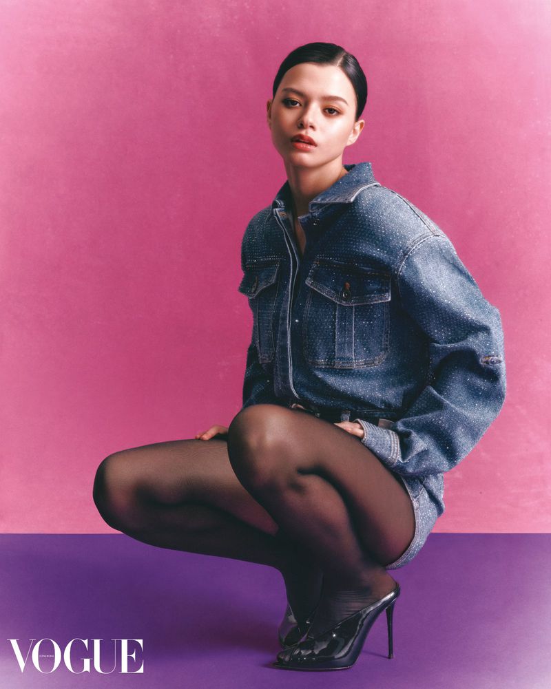 A young woman poses in a denim shirt and tights, kneeling on a purple surface against a vibrant pink background in a studio shot.