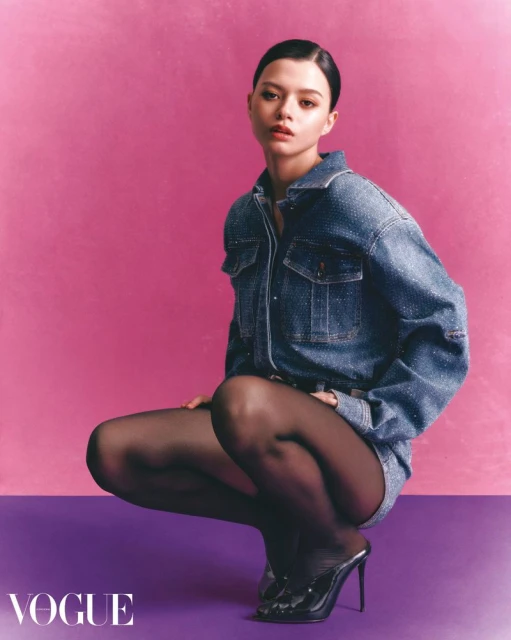 A young woman poses in a denim shirt and tights, kneeling on a purple surface against a vibrant pink background in a studio shot.