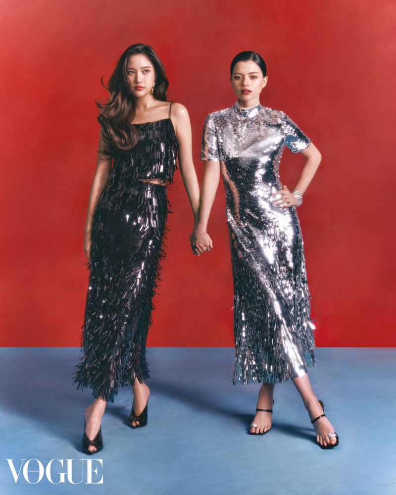 Two women in shimmering dresses hold hands against a red background, showcased in a full shot highlighting fashion and elegant style.