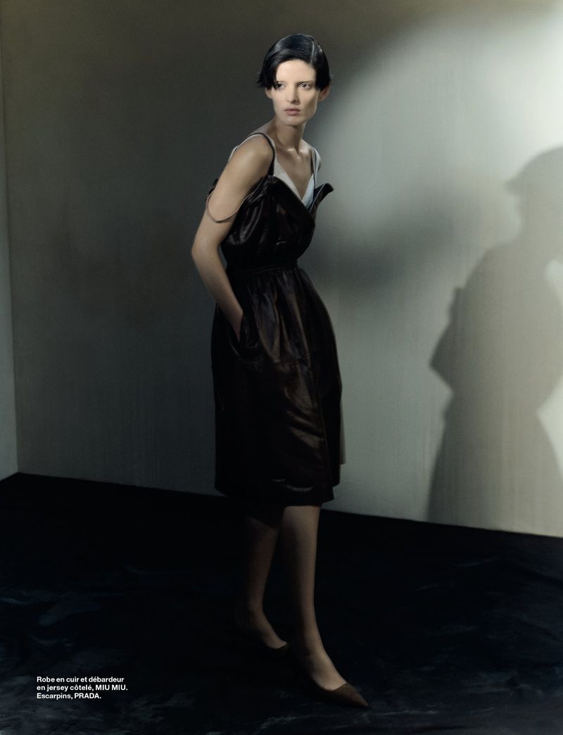 Woman in a dark dress posing indoors against a wall with a shadow, wearing a dress and heels, with a serious facial expression.