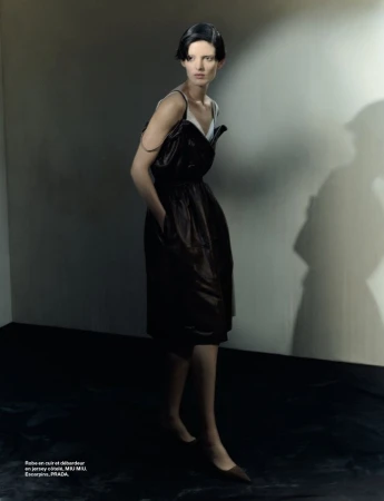 Woman in a dark dress posing indoors against a wall with a shadow, wearing a dress and heels, with a serious facial expression.