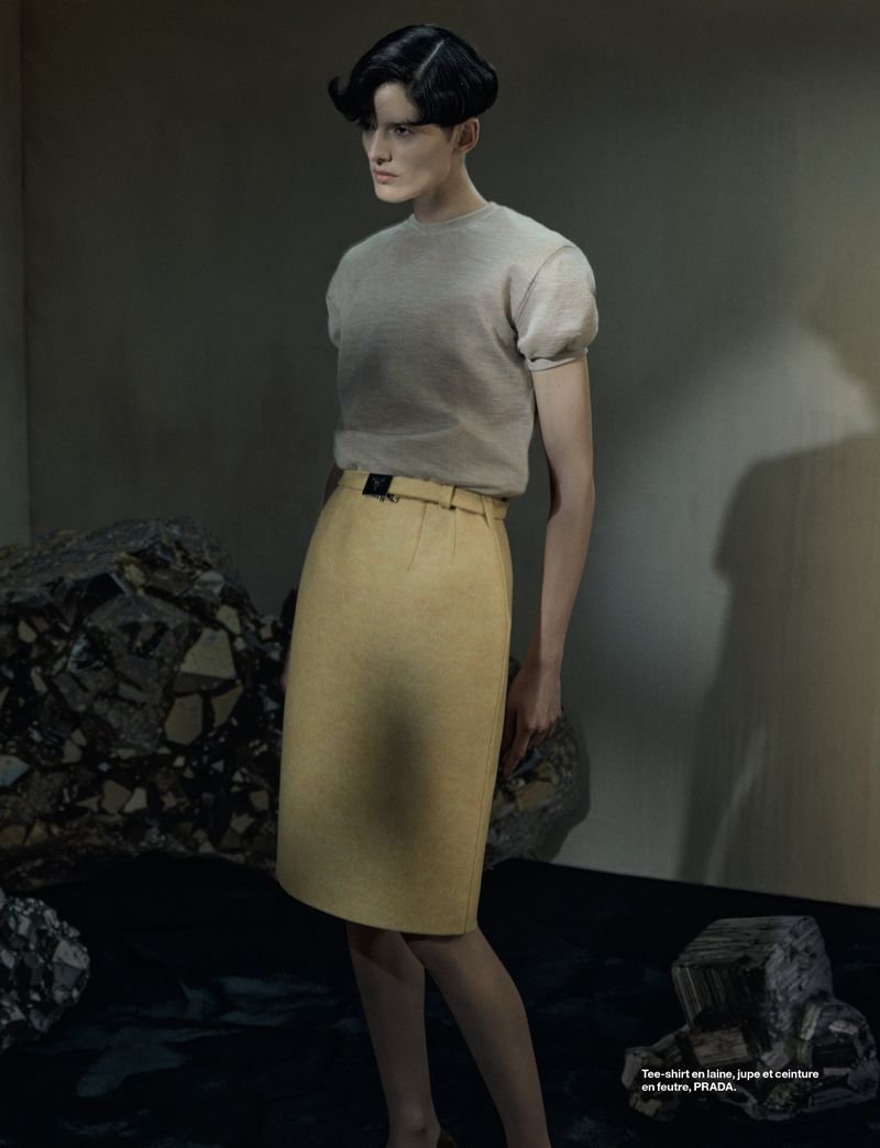 Editorial fashion shot of a slender female model in a pale top and yellow pencil skirt, posing against a mottled grey and black background.