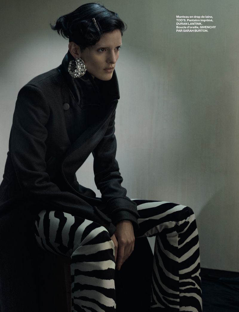 Fashion editorial shot of a woman wearing a black coat and zebra print pants, seated indoors with dramatic lighting and large earrings.