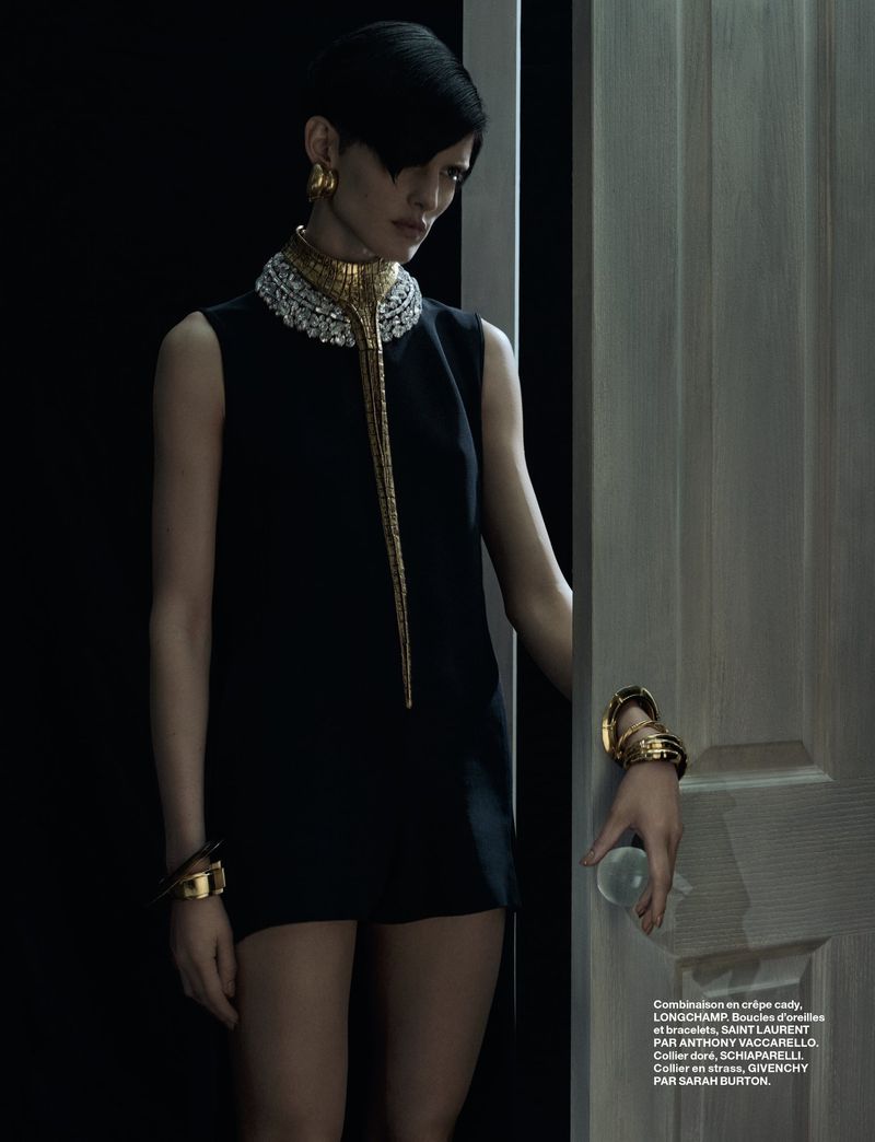 Elegant woman in a short black dress with gold jewelry stands near a doorway in a fashion shot with dark background.