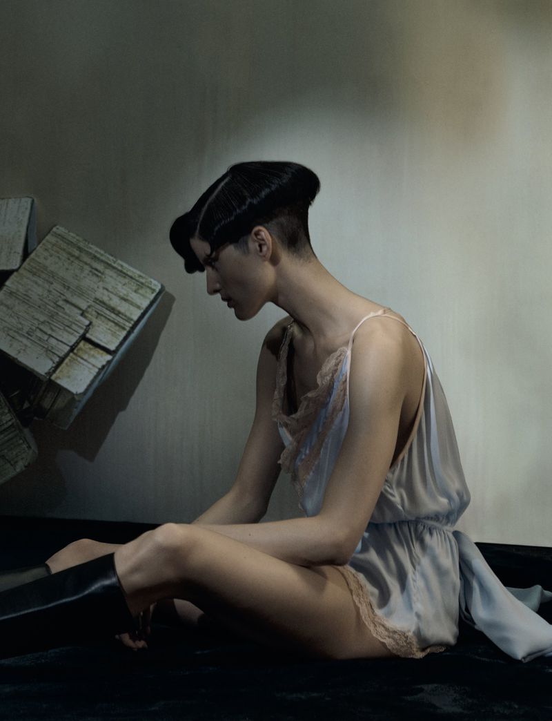 Young woman with short black hair sits with a cubic stone sculpture on a black surface in front of a gray wall, wearing a nightgown.