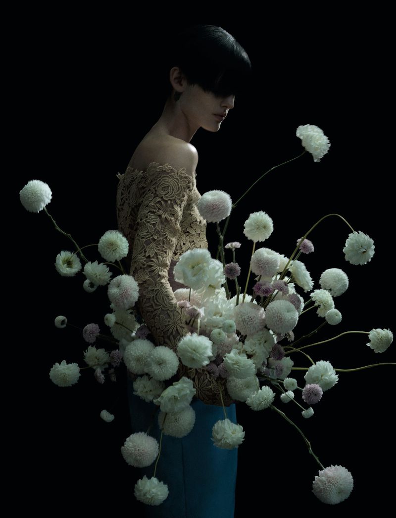 Woman in beige lace top surrounded by white pompom chrysanthemums against a black background in a studio fashion shot.