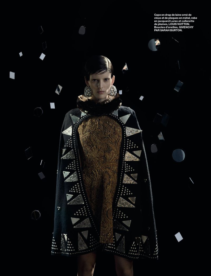 A woman in a gold dress and black, studded cape poses against a dark background with floating geometric metallic shapes.