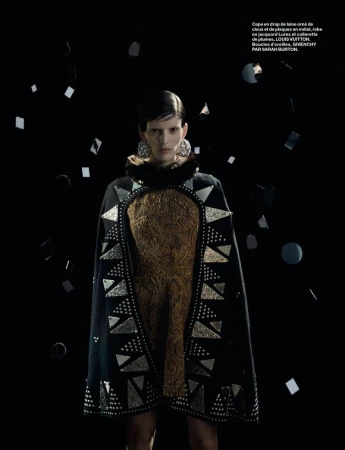 A woman in a gold dress and black, studded cape poses against a dark background with floating geometric metallic shapes.