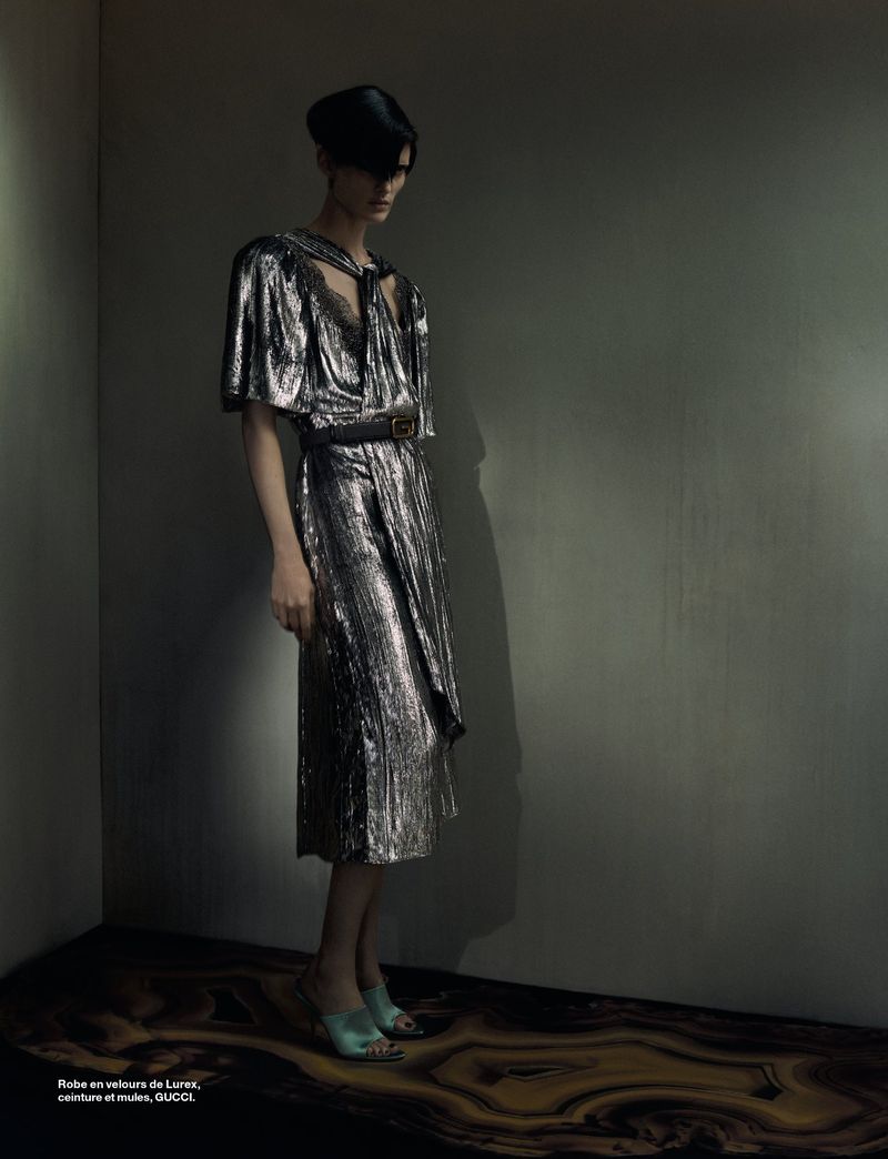 Model in a metallic silver dress and black belt standing against a dark wall for Numéro France