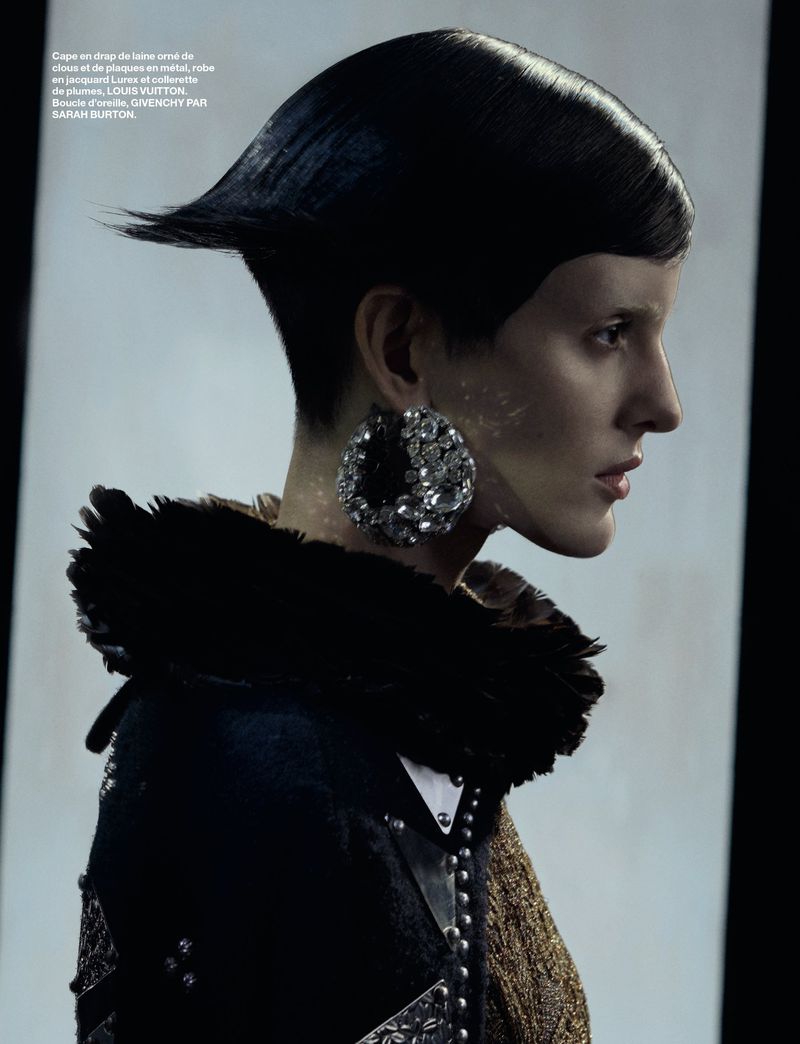 Fashion shot of a woman in a dark wool coat with oversized jewelry, looking to the right in front of a bright, minimalist background.