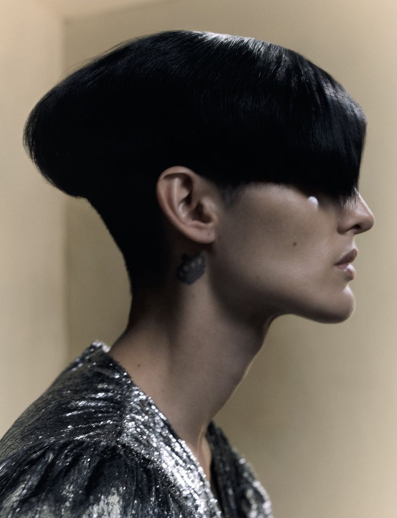 Profile portrait of a person with a short, sleek black hairstyle and silver sequined top against a neutral background in a close-up.
