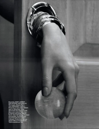 Monochrome close-up of a hand wearing a silver cuff bracelet, grasping a round wooden knob against a wooden door panel.