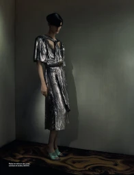 Model in a metallic silver dress and black belt standing against a dark wall for Numéro France