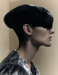 Side profile of model Melany Rivero with a sculptural black bowl haircut wearing a shimmering silver metallic top against a beige wall.