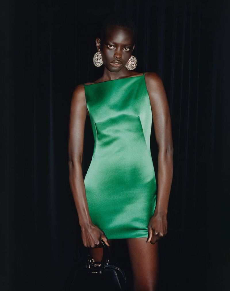 Woman in a green dress and earrings poses in front of dark curtains while holding a black purse in a fashion style photograph.