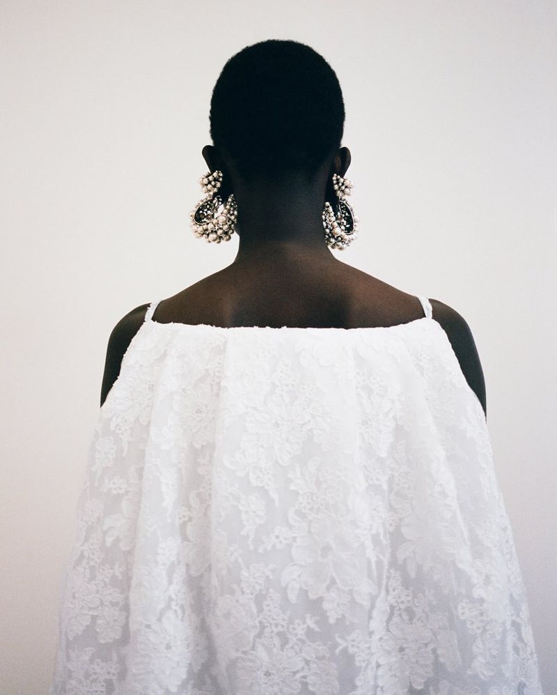 Dark-skinned model with short hair wears white lace dress and large ornate earrings, shot from behind against a plain white background.