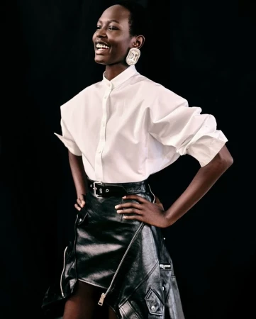 A dark-skinned female fashion model in a white button-up shirt and black leather skirt smiles with hands on hips against a black background.