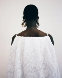 Back view of a model with short cropped hair wearing an airy white lace top and large clustered pearl earrings against a pale background.