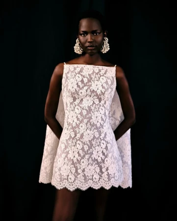 Model wearing a white floral lace mini dress and large pearl cluster earrings against a dark seamless backdrop.