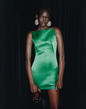 Model in a green satin minidress wearing large round statement earrings against a dark curtain background