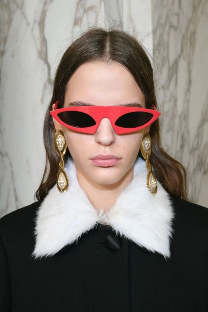 Headshot of a woman with long brown hair wearing red sunglasses, large gold earrings, and a black jacket with a white fur collar.
