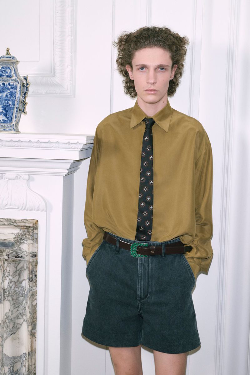 Young adult poses in front of a white mantle wearing a yellow shirt, black tie, jean shorts, and a dark brown belt with green buckle.