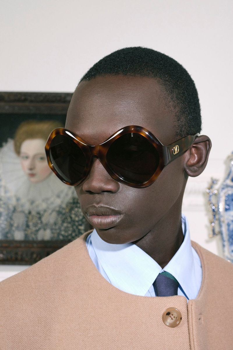 A dark-skinned male model wearing brown geometric sunglasses, a light-blue collared shirt, and a beige jacket in an indoor setting.