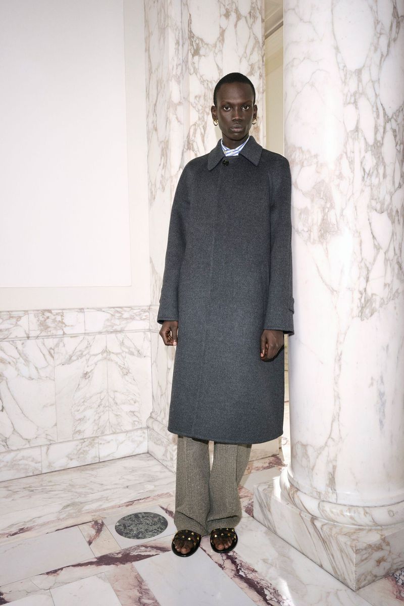 An adult Black male fashion model stands in a grey coat, striped shirt, and patterned trousers near marble columns indoors.