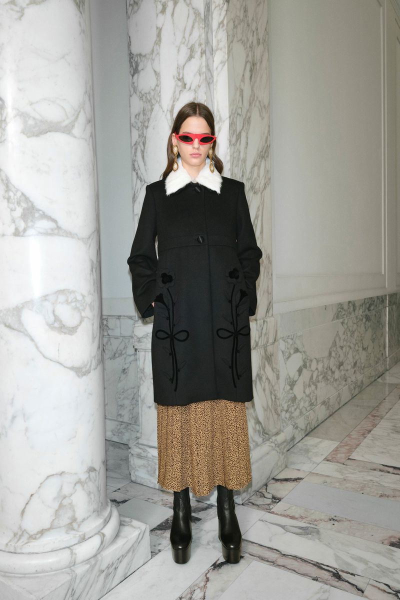 A stylish young woman poses in a black coat and patterned dress against a marble wall backdrop, showcasing modern fashion.